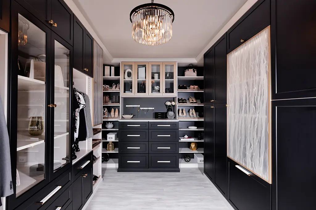 Black shaker walk-in closet with glass-door cabinets, drawer island wall and crystal chandelier