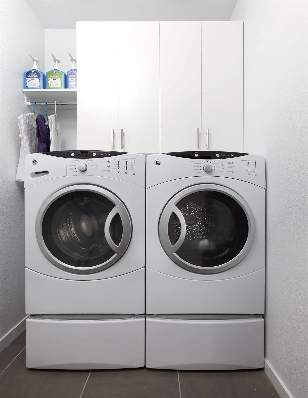 White flat-panel laundry cabinets above a front-load washer and dryer with a hanging rod by Famille Closets